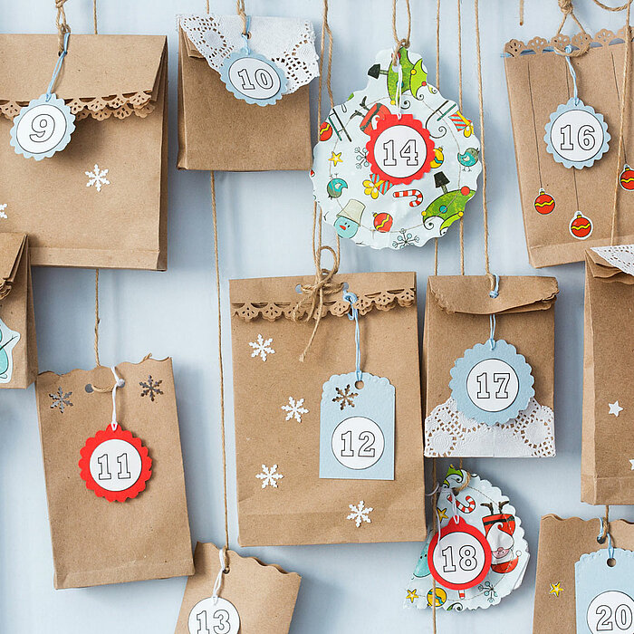 Vierundzwanzig kleine Papiertüten und Umschläge aus braunem Kraftpapier, verziert mit nummerierten Anhängern, Spitzen und weihnachtlichen Motiven, sind an einem hellen Hintergrund aufgehängt. Die nummerierten Anhänger reichen von 1 bis 24, was auf einen A