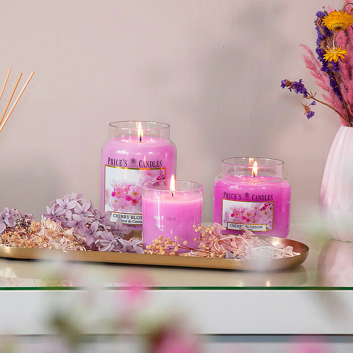 Ein einzelnes, brennendes Duftkerzenglas von Price's Candles mit der Aufschrift "Sandalwood". Die Kerze steht auf einem metallenen Tablett, dekoriert mit Moos und getrockneten Pflanzen. Im Hintergrund ist eine Vase mit Blumen zu sehen, das gesamte Setting
