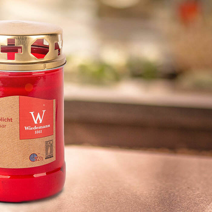 Vor leuchtend gelben Blumen auf einem Steinboden stehen Produkte des Herstellers "Wiedemann". Ein rotes Glasgrablicht mit einer goldfarbenen Abdeckung ist neben einer braunen Verpackung mit der Aufschrift "2 Glasgrablicht Nachfüller" platziert. Auf der Ve