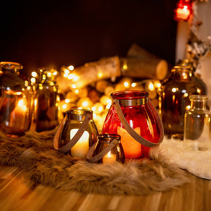 Verschiedene dekorative Glasvasen und Windlichter des Herstellers "Sandra Rich" in unterschiedlichen Größen und Farben, beleuchtet von Kerzenlicht und umgeben von Lichterketten, stehen auf einem gemusterten Holzboden. Die Vasen und Windlichter sind auf we