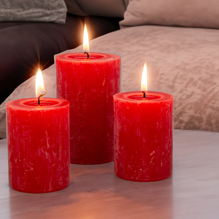 Drei brennende "DecoLite" Kerzen in den Farben Rosa, Rot und Gelb stehen auf einer rustikalen Holzscheibe. Neben den Kerzen sind pinke und weiße Tulpen drapiert. Alles ist auf einem hellen Holztisch arrangiert, was eine gemütliche Frühlingsatmosphäre erze