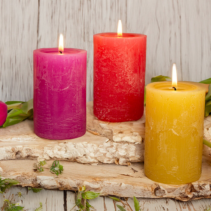 Drei brennende Bolsius Rustikkerzen in verschiedenen Größen in einem sanften Beige. Die Kerzen stehen auf Holzblöcken vor einer Holzwand, umgeben von weißen Blumen, dunklen Steinen, einer gerollten weißen Handtuchrolle und einem kleinen Glasgefäß.