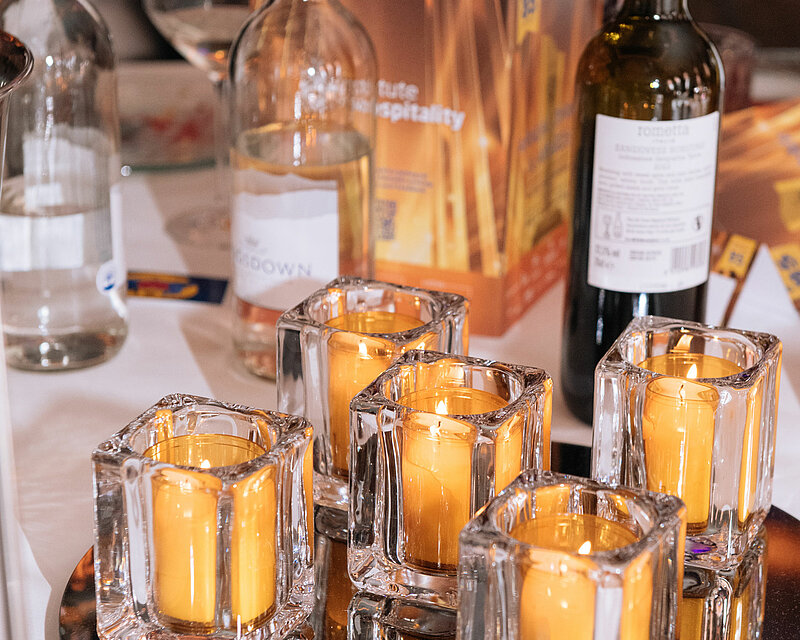 Auf einem festlich gedeckten Tisch stehen Weingläser und Weinflaschen bereit. Ein runder Spiegeluntersetzer liegt auf dem Tisch. Darauf sind mehrere Relight-Halter 'Square' platziert, jeweils mit einem Relight-Nachfüller in Bernsteinfarbe. Die Nachfüller verbreiten ein romantisches und angenehmes Kerzenlicht.