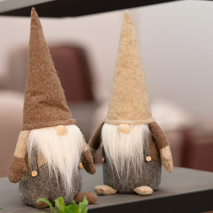 Zwei dekorative Weihnachtswichtel vom Hersteller Edelman stehen auf einem Regal. Sie haben lange weiße Bärte und spitze Hüte, einer in Braun und der andere in Beige. Ihre Körper sind aus grauem Filz gefertigt und sie tragen kleine Knöpfe. Im Vordergrund i