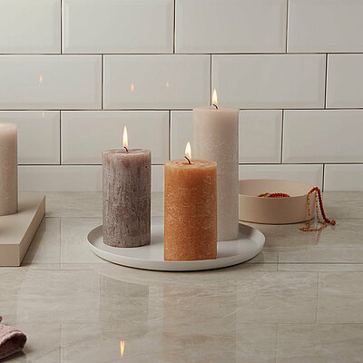 Verschiedene brennende Rustik Stumpenkerzen verschiedener Größe in Farben wie Braun, Weiß, Orange und Anthrazit. Die Kerzen stehen auf weißen Tabletts, vor einer weiß gefließten Wand.