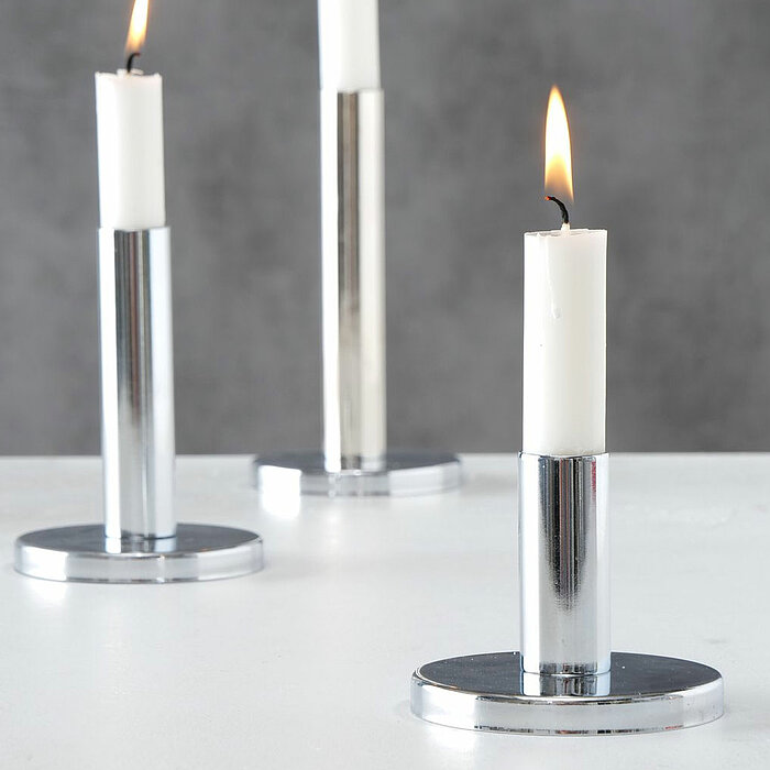 Drei glänzende silberne Kerzenhalter in unterschiedlicher Höhe auf einer Oberfläche vor einem hellen Hintergrund. Daneben steht eine Verpackung mit der Aufschrift "Boltze Home", die ein Bild der Kerzenhalter mit weißen Kerzen zeigt.