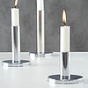 Drei glänzende silberne Kerzenhalter in unterschiedlicher Höhe auf einer Oberfläche vor einem hellen Hintergrund. Daneben steht eine Verpackung mit der Aufschrift "Boltze Home", die ein Bild der Kerzenhalter mit weißen Kerzen zeigt.