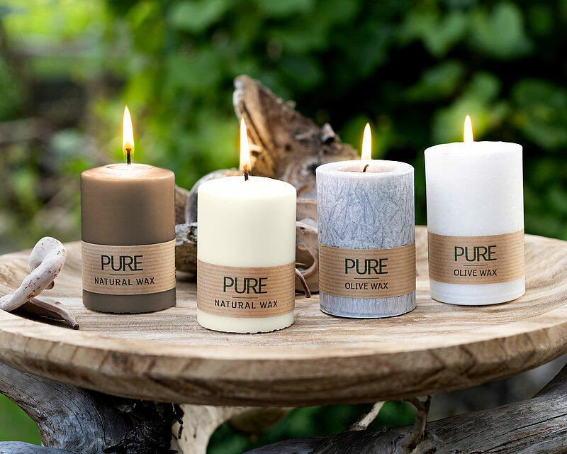 Vier brennende Kerzen aus nachhaltigem Wachs. Zwei davon sind Wenzel Pure Stumpenkerzen in den Farben Sand und Natural. Daneben stehen zwei Wenzel Pure Olivewachs Stumpenkerzen in den Farben Anthrazit und Natur. Die Kerzen stehen auf einem großen hellbraunen Holzteller welcher auf einem dicken Baum Ast steht. Im Hintergrund sieht man verschwommen lichtdurchflutete grüne Blätter.