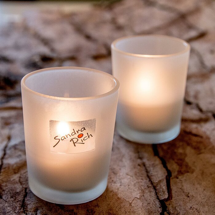 Drei brennende Teelichter in klaren und mattierten Glasbehältern vom Hersteller "Sandra Rich" stehen auf einem runden, schwarzen Tablett mit Griffen. Die schlichten, eleganten Teelichter schaffen eine gemütliche Atmosphäre.