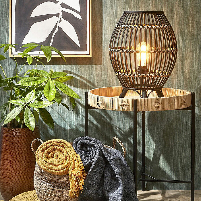 Das Bild zeigt zwei grüne Zimmerpflanzen in strukturierten, braunen Pflanzgefäßen, die auf einem Holzboden stehen. Im Hintergrund liegen zusammengerollte graue Decken in einem geflochtenen Korb, der neben einem gelben Kissen mit grauer Umrandung steht. An