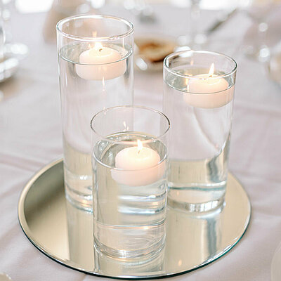 Das gesamte Bild ist in hellen weißen und cremefarbenen freundlichen Tönen. Auf einer weißen Tischdecke wurde festlich gedeckt. In der Mitte des Bildes befindet sich eine schöne runde Spiegelplatte auf welcher sich drei längliche Glasgefäße befinden gefüllt mit Wasser. In jedem Glasgefäß schwimmt jeweils eine brennende weiße Schwimmkerze und es wird eine festliche und gemütliche Stimmung verbreitet.