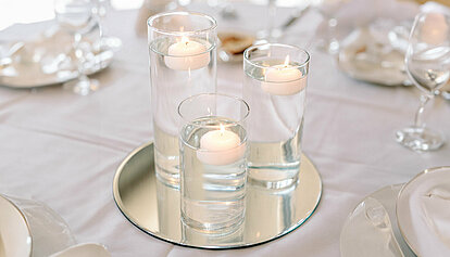 Das gesamte Bild ist in hellen weißen und cremefarbenen freundlichen Tönen. Auf einer weißen Tischdecke wurde festlich gedeckt. In der Mitte des Bildes befindet sich eine schöne runde Spiegelplatte auf welcher sich drei längliche Glasgefäße befinden gefüllt mit Wasser. In jedem Glasgefäß schwimmt jeweils eine brennende weiße Schwimmkerze und es wird eine festliche und gemütliche Stimmung verbreitet.