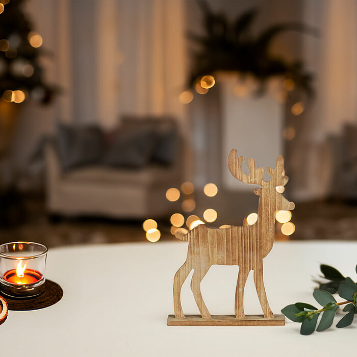 Ein gemütliches Wohnzimmer mit weihnachtlichem Flair. Im Vordergrund steht ein Tisch, auf dem ein Teelicht in einem kleinen Glaswindlicht, getrocknete Orangenscheiben und ein hölzerner Hirsch aufgestellt sind. Daneben liegen einige grüne Zweige. Im Hinter