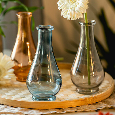 Drei tropfenförmige Deko-Vasen in blau, gelb und orange stehen auf einem geflochtenen Kerzenteller, zusammen mit cremefarbenen Gerbera. Im Hintergrund sieht man verschiedene Topfpflanzen in dekorativen Blumentöpfen aus Kupfer.