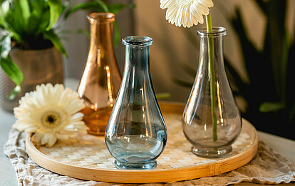 Drei tropfenförmige Deko-Vasen in blau, gelb und orange stehen auf einem geflochtenen Kerzenteller, zusammen mit cremefarbenen Gerbera. Im Hintergrund sieht man verschiedene Topfpflanzen in dekorativen Blumentöpfen aus Kupfer.