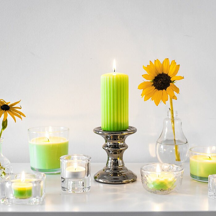 Bolsius Duftkerzen in verschiedenen Formaten und Behältern, hauptsächlich in Grün und Weiß, sind auf einem weißen Tisch dekorativ angeordnet. Eine große grüne Duftkerze steht auf einem silberfarbenen Kerzenhalter in der Mitte. Ringsherum sind kleinere Ker
