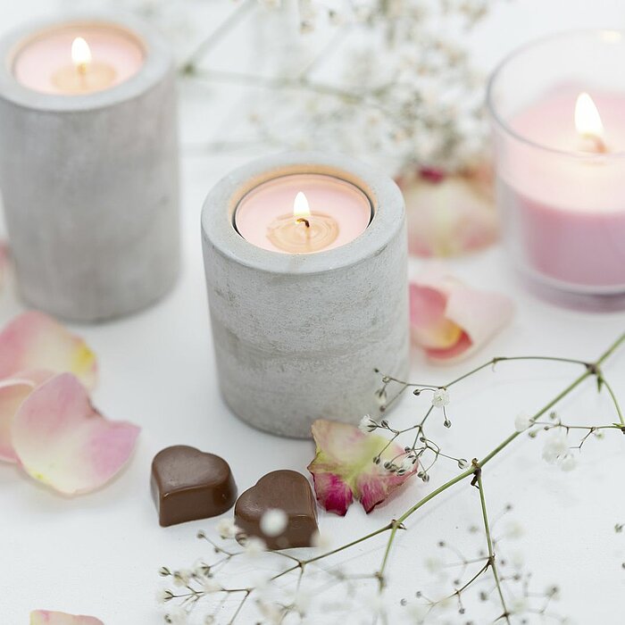 Drei brennende Kerzen von Müller, zwei in zylindrischen Betonhaltern und eine in einem runden Glas, umgeben von Streublumen und Blütenblättern auf einer weißen Oberfläche. Dazwischen liegen zwei kleine, herzförmige Schokoladen. Im Bild sind auch zarte wei