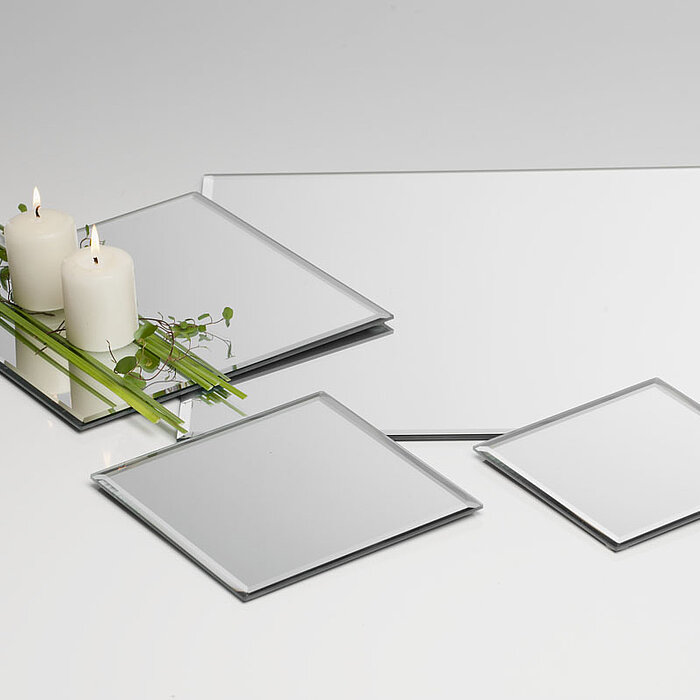 Drei weiße brennende Stumpenkerzen stehen zusammen auf einem quadratischen Spiegel, der mit grünen Blättern und Gräsern dekoriert ist. Um diese Anordnung herum liegen drei weitere rechteckige und quadratische Spiegel unterschiedlicher Größen auf einer wei