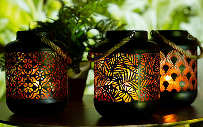 Drei schwarze Metall Laternen mit orientalischen Ausstanzungen, stehen vor einem grün bepflanzten Hintergrund. Durch die ausgestanzten Motive kann man das goldene Innenleben und das warm flackernde Kerzenlicht erkennen.