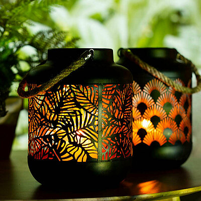 Drei schwarze Metall Laternen mit orientalischen Ausstanzungen, stehen vor einem grün bepflanzten Hintergrund. Durch die ausgestanzten Motive kann man das goldene Innenleben und das warm flackernde Kerzenlicht erkennen.