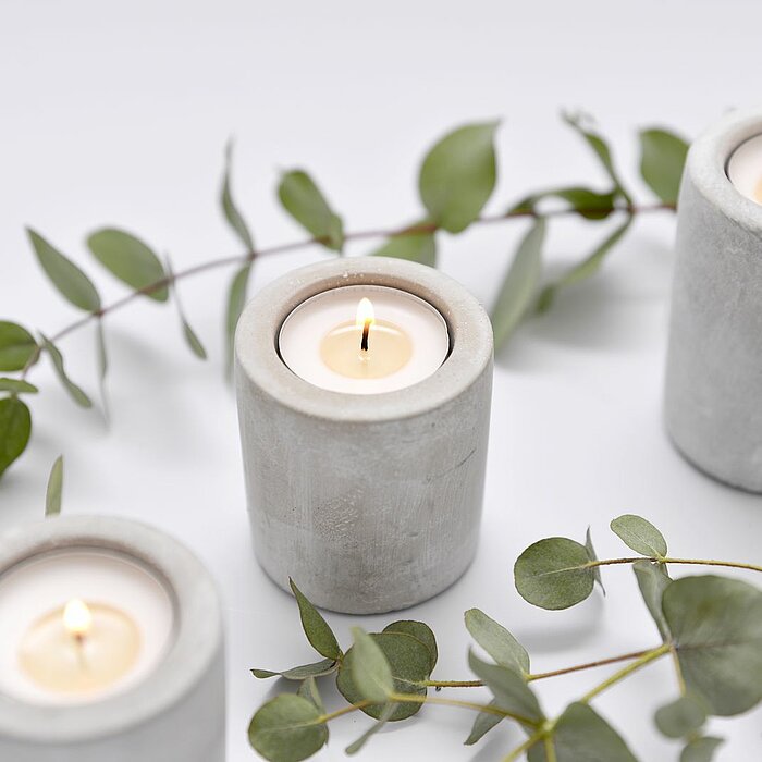 Vier brennende Kerzen des Herstellers "Müller" stehen auf einem hölzernen Tisch. Zwei der Kerzen sind in grauen, rustikalen Haltern, während die anderen beiden in klaren Glashaltern untergebracht sind. Im Hintergrund sind blühende Blumen und eine weiße Te