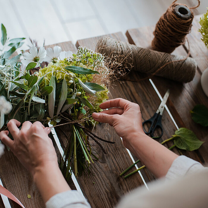 Ein Paar Hände arrangiert ein Bouquet aus verschiedenen Blumen und grünen Zweigen auf einem rustikalen Holztisch. Auf dem Tisch liegt eine Schere, Garnrollen und lose Blätter. Die Szene vermittelt ein Gefühl von Kreativität und handwerklichem Geschick.