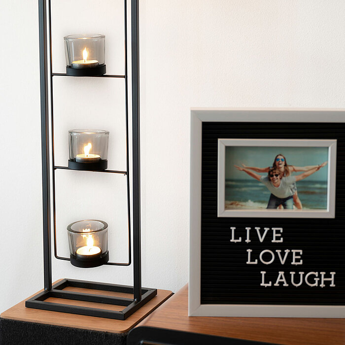 Drei brennende Teelichtkerzen befinden sich in einem modernen, schwarzen Kerzenhalter, der in die Höhe gebaut ist. Daneben steht ein Bilderrahmen auf einem Holztisch, der ein Foto von zwei Personen am Strand zeigt. Unter dem Foto steht der Text "LIVE LOVE