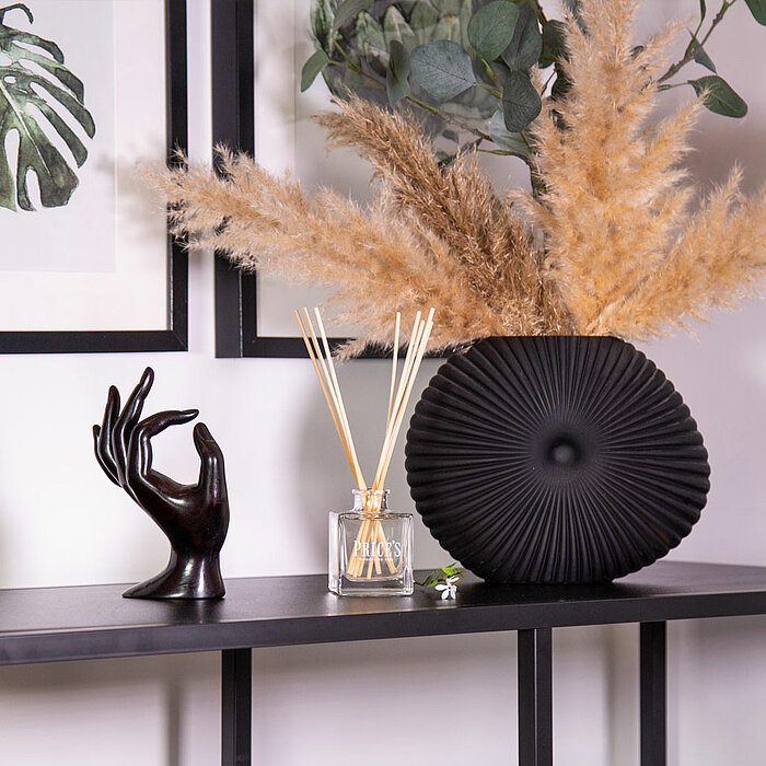 Ein eleganter Konsolentisch mit einer schwarzen Keramikvase, die getrocknete Pampasgraszweige enthält. Daneben steht ein Glasgefäß mit Duftstäbchen von "Prices Candles". Eine schwarze Skulptur einer Hand, die ein stilisiertes Gestenmotiv formt, ist ebenfa