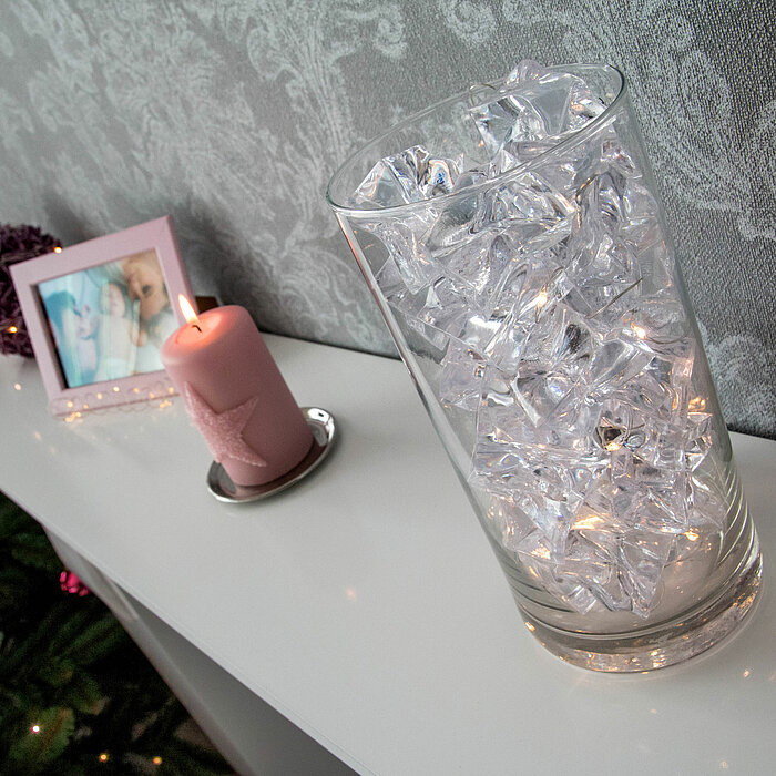 Auf einer weißen Oberfläche steht eine große Glasvase, die mit klaren, funkelnden Eiskristallen und einer Lichterkette gefüllt ist. Daneben befindet sich eine brennende rosa Stumpenkerze mit einem dekorativen Stern auf der Vorderseite, platziert auf einem