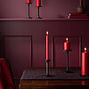 Vier brennende Bolsius Haushaltskerzen in der Farbe Rot in verschiedenen Größen und Formen stehen auf einem dunkelbraunen Tisch. Die Kerzen sind auf schwarzen Kerzenhaltern platziert. Im Hintergrund ist eine weinrote Wand mit dekorativen Elementen und ein
