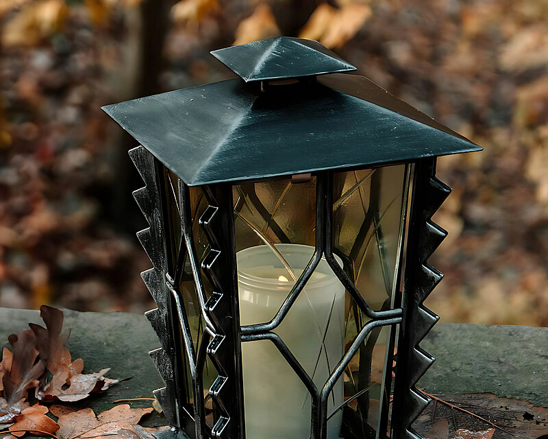 Eine schwarze Stahl-Grablampe in Gussoptik steht auf einer Mauer, umgeben von leicht gefrorenem Herbstlaub. Im Hintergrund ist ein schöner Herbstwald zu erkennen.