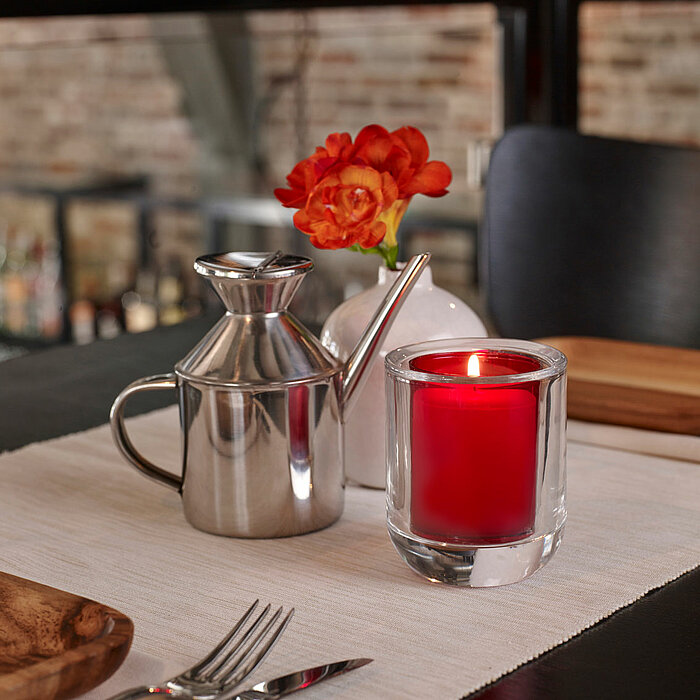 Zwei Bolsius Votivkerzen in quadratischen Glasbehältern stehen auf einem Holz-Esstisch. Neben den Kerzen befindet sich eine weiße Vase mit einer rosa Orchidee. Im Hintergrund sind mehrere Holztische und weiße Stühle mit einem Filigranmuster zu sehen.