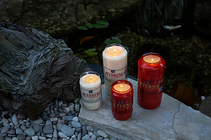 In einem Steinbeet liegt ein flacher großer grauer Stein auf welchem zwei weisse und zwei rote brennende Aeterna Grabkerzen platziert sind. Im Hintergrund ist ein schöner Garten zu sehen.