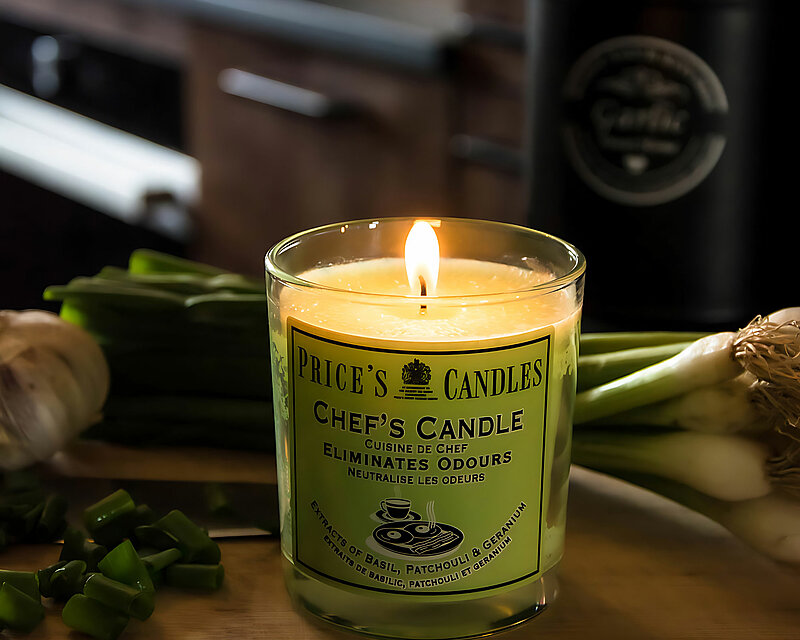 Ein grünes Duftglas Chefs Candle von Prices steht auf einer Holzarbeitsplatte in einer dunkelbraunen Küche. Auf der Holzarbeitsplatte befinden t sich ein Bund Frühlingszwiebeln und ein paar geschnittene Frühlingszwiebeln und ein Knoblauch.