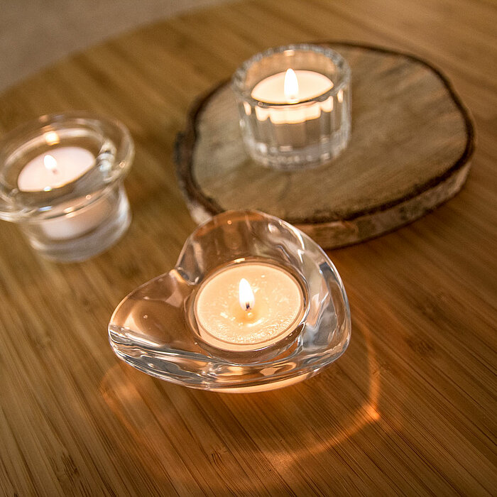 Eine brennende Kerze in einem klaren, geriffelten Glasbehälter des Herstellers "Sandra Rich" steht auf einer dekorativen Holzscheibe. Das warme Licht der Kerze wirft sanfte, strahlenförmige Muster auf die Holzunterlage.