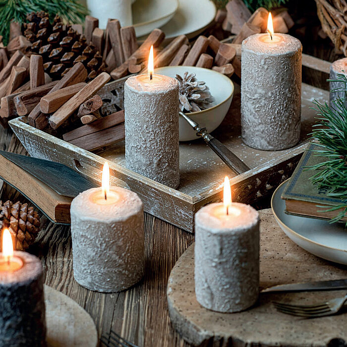Sechs brennende, rustikale Stumpenkerzen des Herstellers "Wenzel" in verschiedenen Größen und gedecktem Creme-Ton stehen auf einem rustikalen Holztisch, der mit Tannenzapfen, Holzstücken und einem Stapel Bücher dekoriert ist. Auf einem weißen Teller liegt