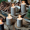 Sechs brennende, rustikale Stumpenkerzen des Herstellers "Wenzel" in verschiedenen Größen und gedecktem Creme-Ton stehen auf einem rustikalen Holztisch, der mit Tannenzapfen, Holzstücken und einem Stapel Bücher dekoriert ist. Auf einem weißen Teller liegt