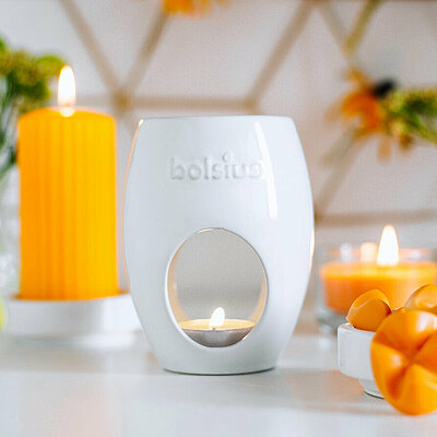 Auf dem Bild befindet sich eine weisse Duftlampe Oval von Bolsius mit einem brennenden Teelicht. Daneben steht eine Schale mit mehrere orangefarbenen Wachsmelts. Im Hintergrund steht eine gelbe geriffelte brennende orangefarbene Stumpenkerze. Auf dem Bild sind verschiedene Vasen und Glasvasen mit verschiedenen grünen Blumen.