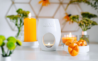 Auf dem Bild befindet sich eine weisse Duftlampe Oval von Bolsius mit einem brennenden Teelicht. Daneben steht eine Schale mit mehrere orangefarbenen Wachsmelts. Im Hintergrund steht eine gelbe geriffelte brennende orangefarbene Stumpenkerze. Auf dem Bild sind verschiedene Vasen und Glasvasen mit verschiedenen grünen Blumen.