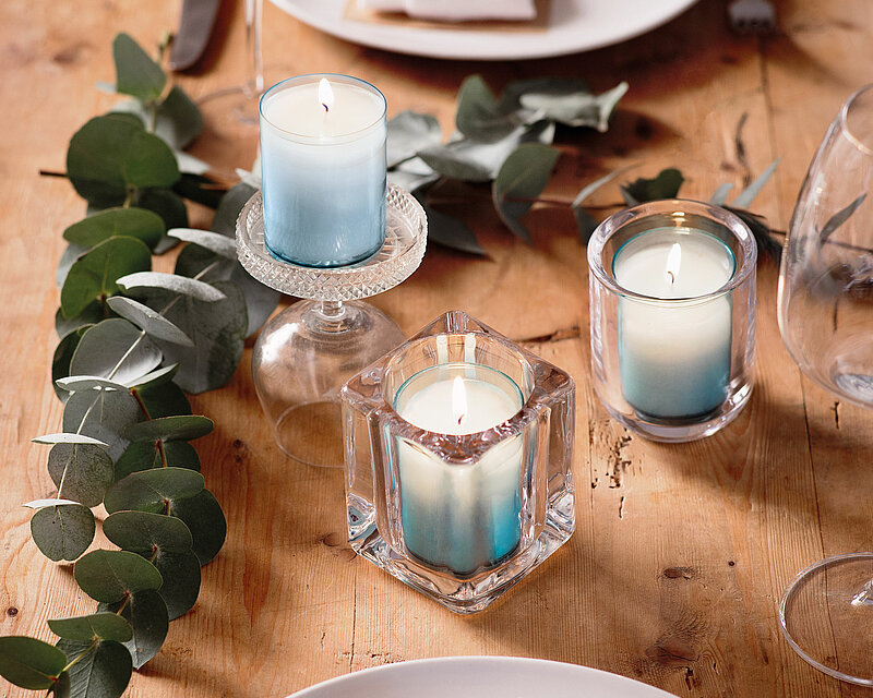 Auf einem gedeckten Holztisch stehen drei unterschiedliche Relight Halter von Bolsius mit je einer brennenden Relight Nachfüllkerze von Bolsius in der Farbe Aqua. Daneben befinden sich einige Blätter was dem gesamten Bild eine warme einladende und festliche Atmosphäre als sitzt man an einem gemütlichen Frühstückstisch verleiht.