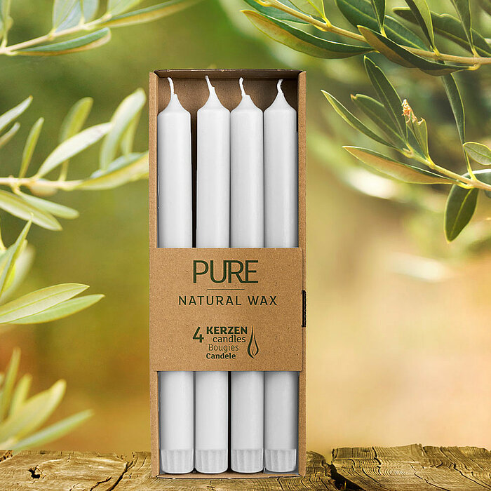 Mehrere Zweige eines Olivenbaums mit grünen Oliven und Blättern ragen in das Bild hinein. Im Vordergrund befindet sich eine rustikale hölzerne Oberfläche, die möglicherweise als Tisch dient. Der Hintergrund ist verschwommen und in warmen, hellen Farben ge