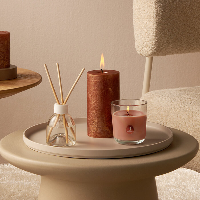 Bolsius Duftkerzen: Auf einem Holztisch befinden sich vier Bolsius Produkte. Links steht eine brennende gold-bronzefarbene Bolsius Duftkerze auf einem modernen weißen Kerzenhalter. Daneben befindet sich ein transparentes Glasfläschchen mit Diffusorsticks.