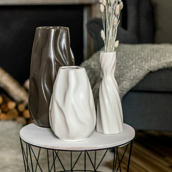 Acht Vasen vom Hersteller "Sandra Rich" in verschiedenen Formen und Erdtönen stehen auf einem beige gedeckten Tisch. Die Vasen enthalten diverse Trockenblumen wie Pampasgras und Schleierkraut. Auf dem Tisch sind kleine Steine und Stofftücher verteilt, was