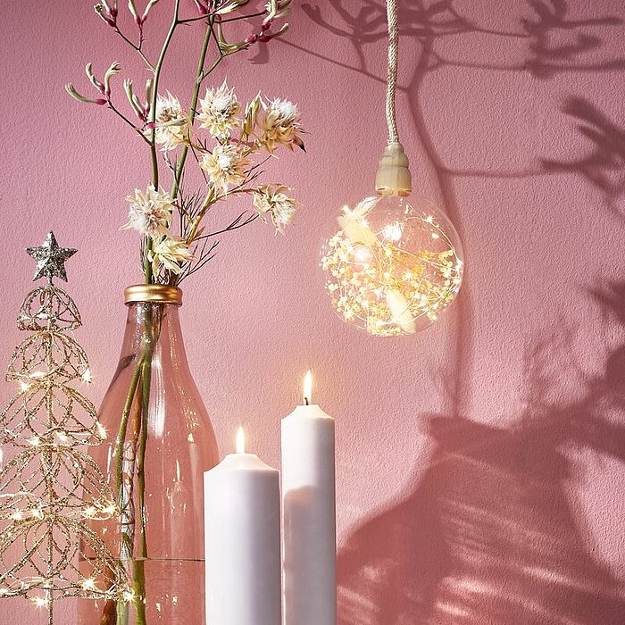 Eine kugelförmige Hängelampe mit einem Durchmesser von 14 cm und einer Höhe von 14 cm hängt an einem 80 cm langen Seil. Das durchscheinende Lampengehäuse enthält filigrane, beleuchtete Pflanzenmuster, die eine warme und gemütliche Atmosphäre ausstrahlen. 