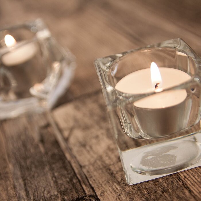 Mehrere kleine Teelichter, platziert in stilvollen, transparenten Glasbehältern des Herstellers Sandra Rich, erleuchten eine dunkle Oberfläche. Im Hintergrund sind weitere brennende Kerzen zu sehen, die eine sanfte und gemütliche Atmosphäre schaffen.