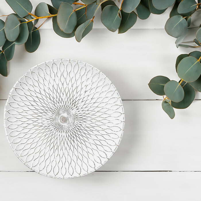 Eine gemütliche Szene in neutralen Tönen. Eine grob gestrickte weiße Decke liegt auf einem Sofa, daneben ein aufgeschlagenes Buch. Auf einem kleinen weißen Tisch, der schräg im Hintergrund steht, befindet sich eine Tasse Kaffee auf einer Servierschale mit