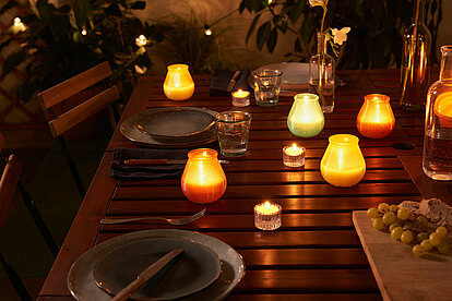 Auf dem Bild ist eine gemütliche Terrasse im Abendlicht zu sehen. Auf einem schön eingedeckten Holztisch stehen unterschiedlich farbene Bolsius True Citronella Patiolichter, die einen wunderbaren hellen Kerzenschein verbreiten. Dazwischen sind mehrere Bolsius True Citronella Teelichter angeordnet, die ebenfalls das Bild mit ihrem warmen Licht erhellen. Im Hintergrund sind verschwommen einige grüne Balkonpflanzen zu sehen, die mit einer hübschen Lichterkette dekoriert sind. Die Szene strahlt ein schönes und gemütliches Ambiente an einem lauen Sommerabend aus.