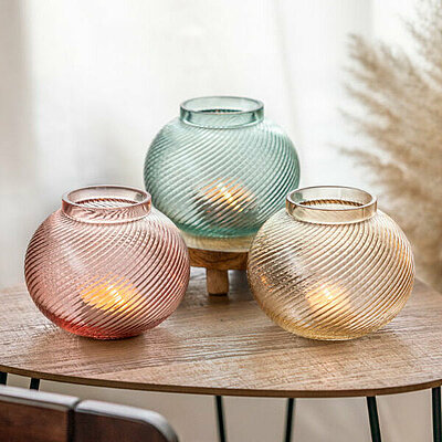 Drei bauchige Kerzenhalter aus buntem Glas, perfekt für deine Frühlingsdeko, stehen auf einem kleinen Holztisch. Im Hintergrund steht eine braune Keramikvase mit buschigem Schilfgras.