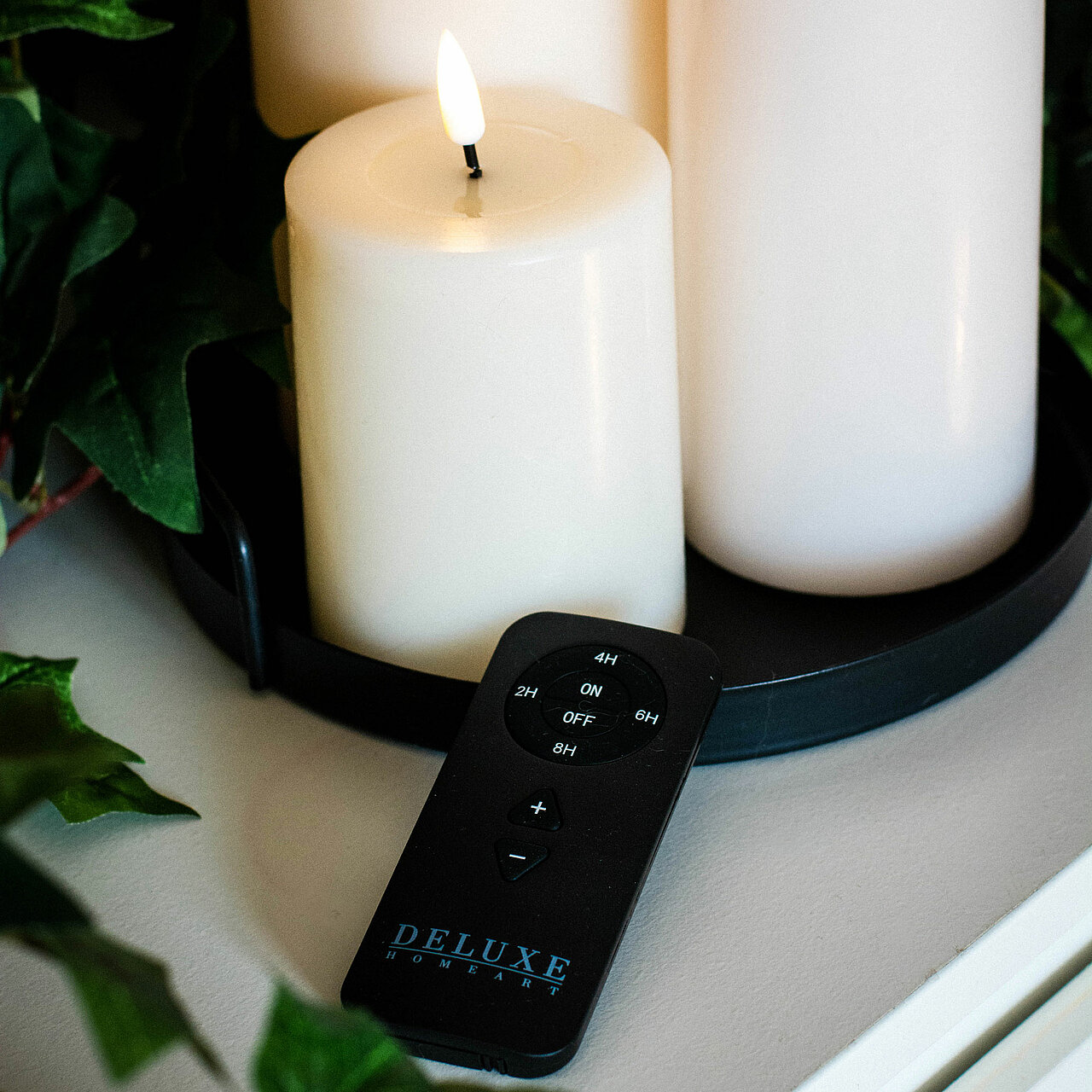 Nahaufnahme einer schwarzen Fernbedienung für LED Kerzen von Deluxe Homeart. Im Hintergrund sind leuchtende LED Stumpenkerzen zu sehen.