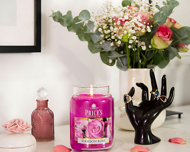 Auf einer weißen Kommode steht eine brennende Duftkerze von Prices im Apothekerglas mit 630 Gramm, gefüllt mit sattem pinken Wachs und dem Duft von Damson Rose. Um das Glas herum sind einige Rosenblüten verteilt. Rechts daneben steht eine schwarze Hand, an deren Fingern goldene Schmuckringe stecken. Im Hintergrund ist eine weiße Vase mit einem bunten Blumenstrauß aus Rosen und weißen Maiglöckchen zu sehen. Die Szene wirkt elegant und stilvoll.